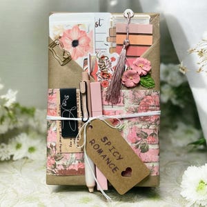 May include: A decorative gift box wrapped in floral paper and tied with twine. The box contains stationery items, including pens, cards, and a tassel. A tag with the words "Spicy Romance" hangs from the box.