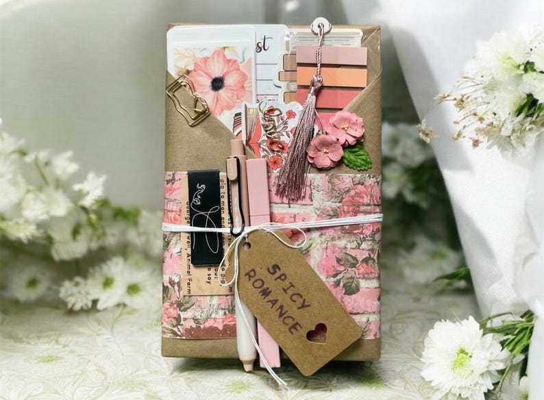 May include: A decorative gift box wrapped in floral paper and tied with twine. The box contains stationery items, including pens, cards, and a tassel. A tag with the words "Spicy Romance" hangs from the box.