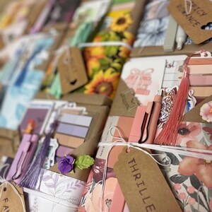 May include: A collection of handmade bookish gifts, including bookmarks, pens, and decorative elements. The items are wrapped in brown paper and tied with twine, featuring tags labeled "Fantasy", "Thriller", and "Spicy Romance". The color palette includes pinks, purples, and floral patterns.