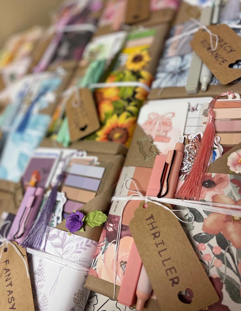 May include: A collection of handmade bookish gifts, including bookmarks, pens, and decorative elements. The items are wrapped in brown paper and tied with twine, featuring tags labeled "Fantasy", "Thriller", and "Spicy Romance". The color palette includes pinks, purples, and floral patterns.
