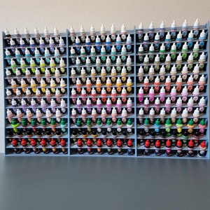 Op de afbeelding: Drie lichtblauwe verfopslagrekken gevuld met talrijke kleine verfflesjes. De flesjes zijn gerangschikt op kleur en tonen een breed spectrum aan tinten, van levendig rood en geel tot koele blauw en groen. De planken zijn netjes georganiseerd.