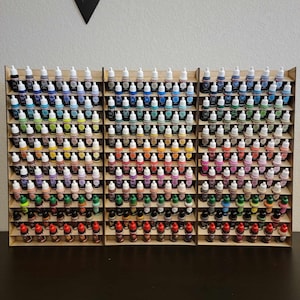 May include: A wooden paint rack with multiple shelves, holding rows of small bottles of paint in various colors. The bottles are arranged in a rainbow pattern, with the lightest colors at the top and the darkest colors at the bottom.