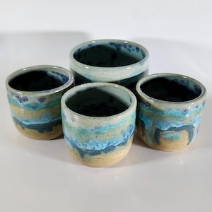 May include: Four ceramic cups with a unique, layered design. The cups feature a blend of colors, including beige, blue, and teal, creating a textured, watercolor effect. The cups vary in size, with one taller than the others.