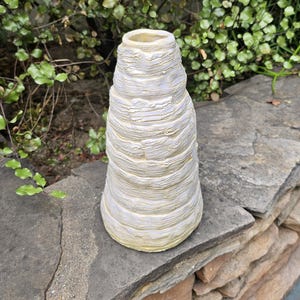 May include: A conical ceramic vase with a textured, layered surface featuring horizontal ridges. The exterior is finished in an off-white glaze with subtle yellow undertones visible within the crevices of the stacked, uneven clay rings.