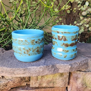 May include: Two turquoise ceramic planters with a textured, speckled design. The planters have a cylindrical shape, with horizontal bands of brown and turquoise. The planters are sitting on a stone surface, with greenery in the background.