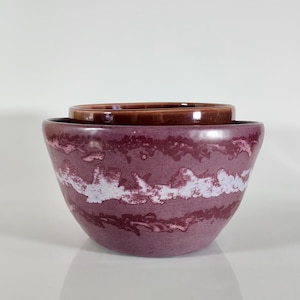 May include: Two ceramic bowls, one nested inside the other. The larger bowl is a deep purple with a white, marbled design. The smaller bowl is a dark brown. The bowls are on a white surface.