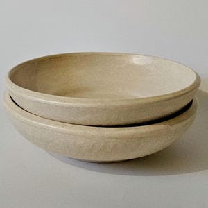 May include: Two beige ceramic bowls stacked on top of each other. The bowls have a simple, round design with a slightly textured surface. The neutral color and minimalist style make them suitable for various uses.
