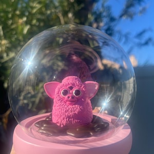 May include: A pink Furby toy inside a clear glass snow globe with a pink base. The Furby is sitting on a bed of brown chocolate sweets.