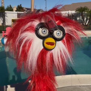 May include: A vintage Furby toy with red and white shaggy fur. The toy has large, round eyes, a yellow beak, and black eye sockets. The Furby is suspended in the air against a bright blue sky and a swimming pool.