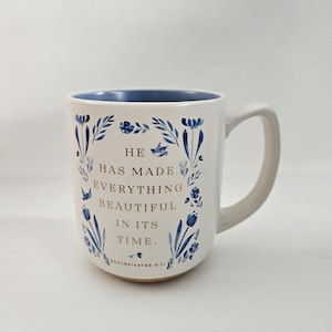 May include: White ceramic mug with a blue interior and a curved handle. The mug is decorated with blue floral designs and the text "HE HAS MADE EVERYTHING BEAUTIFUL IN ITS TIME." The text "Ecclesiastes 3:11" is at the bottom.