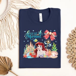 a blue t-shirt with a design of a mermaid surrounded by various sea-themed elements, such as a castle, seashells, and starfish.