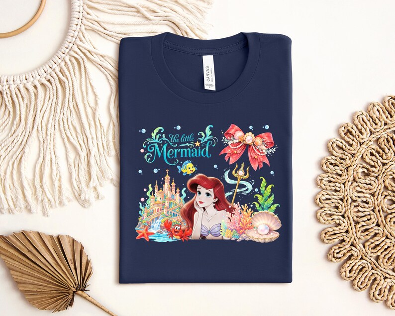 a blue t-shirt with a design of a mermaid surrounded by various sea-themed elements, such as a castle, seashells, and starfish.