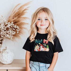 A young girl with blonde hair and blue eyes is smiling and posing in front of a floral arrangement and a black t-shirt with a mermaid design.