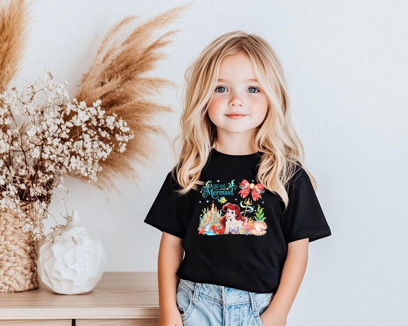 A young girl with blonde hair and blue eyes is smiling and posing in front of a floral arrangement and a black t-shirt with a mermaid design.