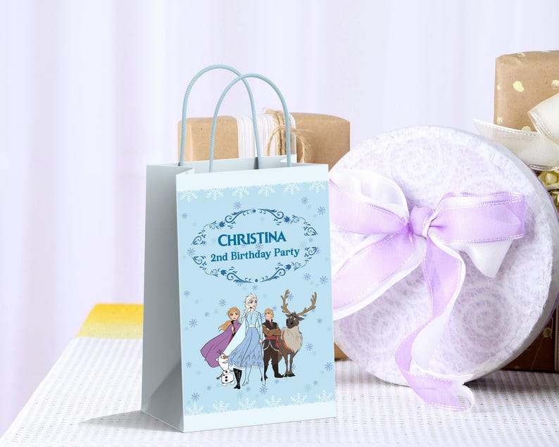 May include: A pale blue party bag featuring characters from the film Frozen, with the text "CHRISTINA 2nd Birthday Party". The bag has light blue handles and is next to a round gift box tied with a lilac ribbon.