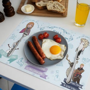 May include: A placemat featuring characters from the movie Frozen, with a plate of food including sausage, a fried egg, and tomatoes. A glass of orange juice and a cutting board with bread are also visible.