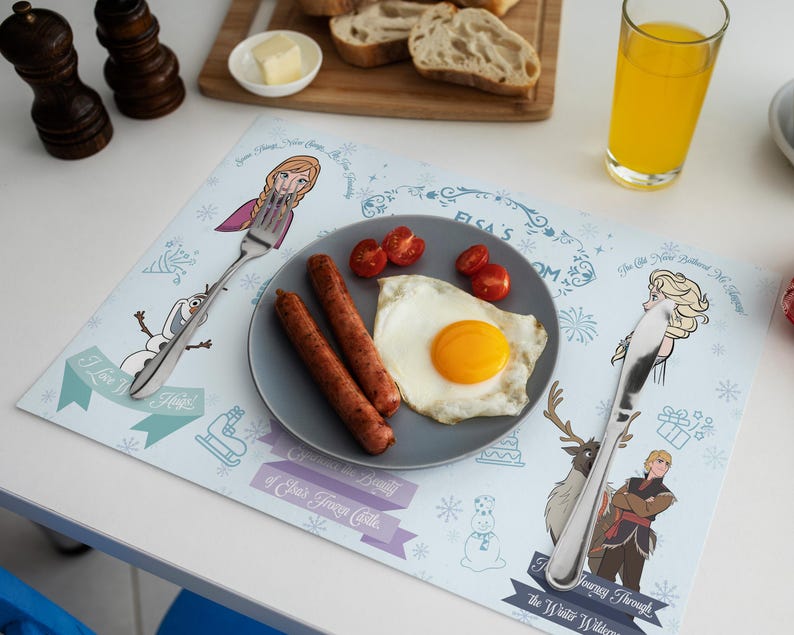 May include: A placemat featuring characters from the movie Frozen, with a plate of food including sausage, a fried egg, and tomatoes. A glass of orange juice and a cutting board with bread are also visible.