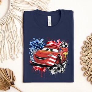 A navy blue t-shirt with a graphic of a red car, featuring an American flag and splatter paint, is displayed on a white background.
