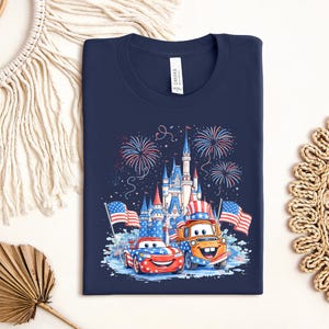 A navy blue t-shirt with a graphic design featuring a castle, fireworks, and two cartoon cars, one orange and one red, along with American flags.