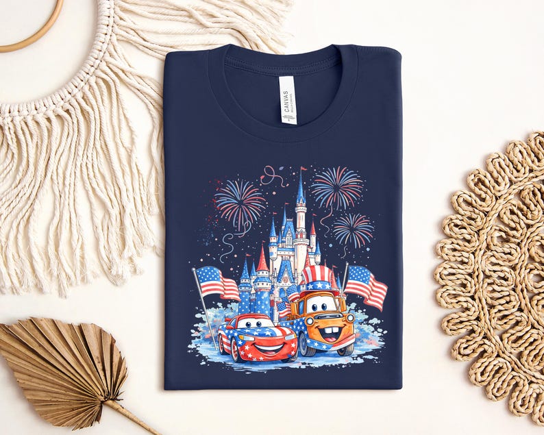 A navy blue t-shirt with a graphic design featuring a castle, fireworks, and two cartoon cars, one orange and one red, along with American flags.