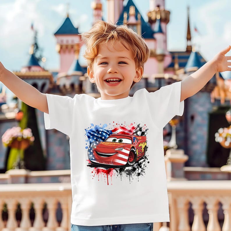 A young boy with blond hair is smiling and raising his arms, wearing a white t-shirt with a graphic of a red car and an American flag.