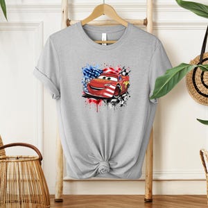 A gray t-shirt with a graphic of a red car and an American flag design, hanging on a wooden hanger.