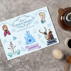 May include: A light blue placemat featuring Elsa's Magical Kingdom theme with illustrations of characters and elements from the movie. The placemat includes text and decorative borders, with a teapot, cup of coffee, and sugar bowl in the background.