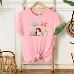 A pink t-shirt with a mermaid design is hanging on a wooden hanger, surrounded by various decorative items such as a woven basket and a potted plant.