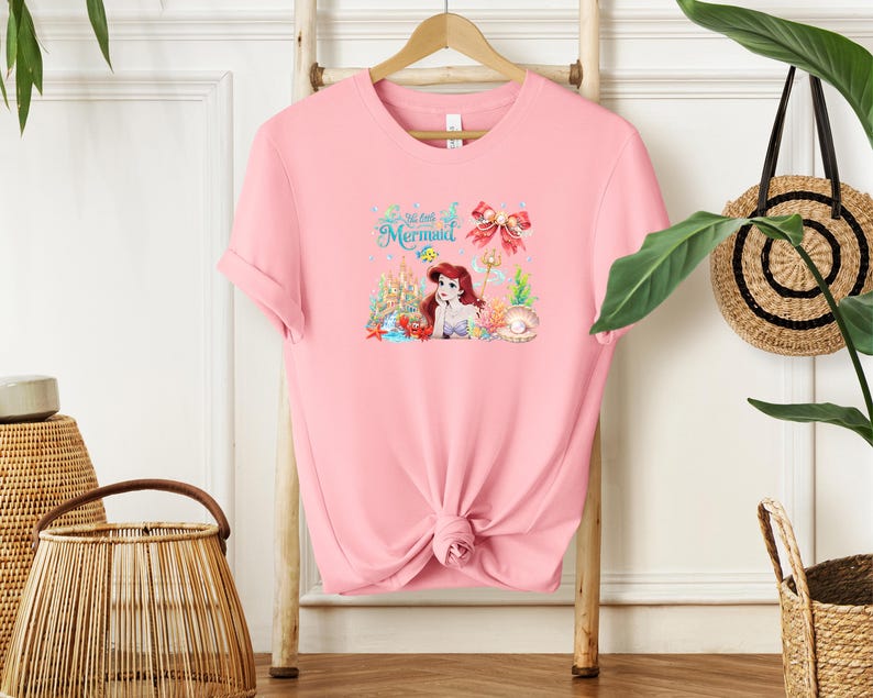A pink t-shirt with a mermaid design is hanging on a wooden hanger, surrounded by various decorative items such as a woven basket and a potted plant.