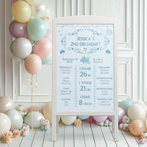 Puede incluir: Un letrero enmarcado en blanco con el texto "JESSICA'S 2ND BIRTHDAY!" en azul claro. El letrero incluye detalles sobre el cumplea&ntilde;os, incluyendo altura (66 cm), peso (9,5 kg) y n&uacute;mero de dientes (8). Globos pastel rodean el letrero.