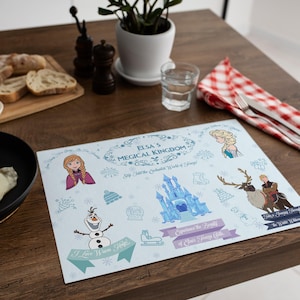 May include: A light blue placemat featuring illustrations of characters and elements from the movie "Frozen." The placemat includes the text "Elsa's Magical Kingdom" and other phrases. The placemat is on a wooden table.