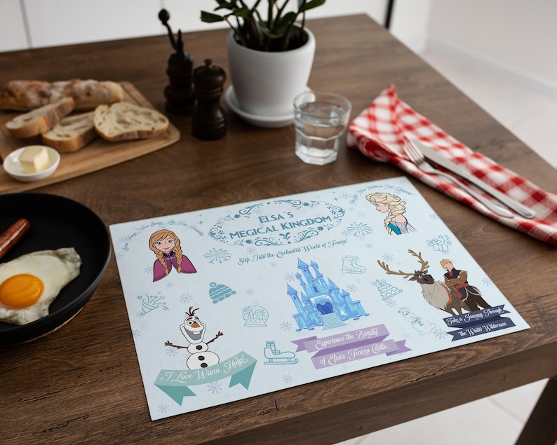 May include: A light blue placemat featuring illustrations of characters and elements from the movie "Frozen." The placemat includes the text "Elsa's Magical Kingdom" and other phrases. The placemat is on a wooden table.