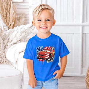 A young boy wearing a blue t-shirt with a graphic of a red car and an American flag design, smiling at the camera.