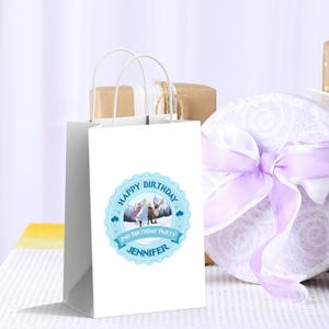 May include: A white gift bag with a blue circular label that reads "Happy Birthday" and "2nd Birthday Party Jennifer". The bag has white handles and is next to a round gift box with a purple ribbon.