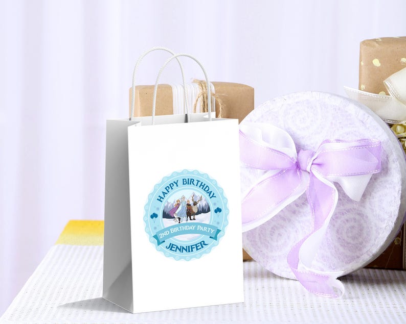May include: A white gift bag with a blue circular label that reads "Happy Birthday" and "2nd Birthday Party Jennifer". The bag has white handles and is next to a round gift box with a purple ribbon.