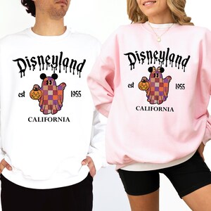 Peut inclure: Un sweat-shirt blanc et un sweat-shirt rose clair, chacun arborant un motif de fantôme avec des oreilles de Mickey tenant une citrouille. Le design inclut le texte Disneyland, est 1955, et California en noir, avec le fantôme dans un motif patchwork violet et orange.