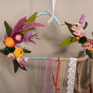 May include: A blue and brown wooden wreath decorated with dried flowers, including pink, purple, yellow, and orange blooms. The wreath is adorned with various lace trims in shades of purple, orange, and white.
