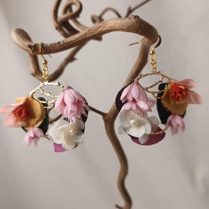 May include: Floral earrings with gold-colored hoops and delicate flower accents in shades of pink, white, and orange. The earrings are displayed on a natural, curved branch, showcasing their intricate design and craftsmanship.