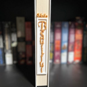May include: A book with a white spine and beige pages. A 3D-printed orange-colored text that reads "Hello Beautiful" is attached to the spine. The book is on a black shelf with other books in the background.