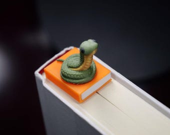 May include: A green and yellow snake figurine rests on a small orange book, positioned on the edge of a larger book. The snake is coiled and has a detailed design. The books are set against a dark background.