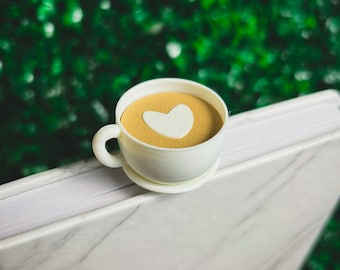 Puede incluir: Una taza de café de cerámica blanca con un platillo, con un diseño de café de color marrón claro con una forma de corazón blanco en el centro. La taza está sobre una superficie de mármol blanco, con un fondo verde borroso.