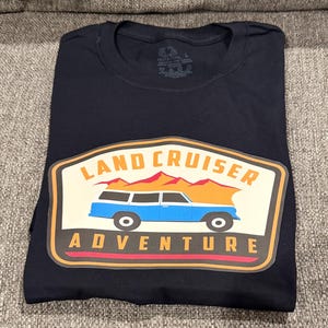May include: A navy blue t-shirt featuring a graphic with the words "LAND CRUISER ADVENTURE". The graphic includes a blue station wagon, orange mountains, and a cream-colored background.