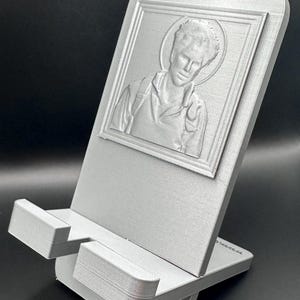 Op de afbeelding: Zilverkleurige telefoonstandaard met een portret van een persoon met een halo. Het portret staat in een rechthoekige lijst. De standaard heeft een voet en een steunarm met een sleuf om een telefoon vast te houden. De standaard is van metaal.