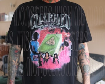 Puede incluir: Camiseta negra con un estampado gráfico del programa de televisión "Charmed". El gráfico presenta el título del programa, un libro con un símbolo triquetra y los personajes del programa. El texto en el gráfico dice "Something Wicca This Way Comes."