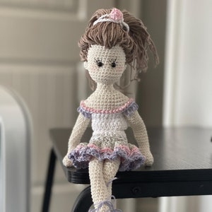 May include: A crocheted doll wearing a white dress with pink and purple trim, brown hair, and purple shoes. The doll is sitting on a black table.