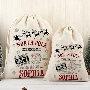May include: Two beige Christmas gift bags with drawstring closures. The bags feature a black silhouette of Santa's sleigh and reindeer, with the text "North Pole Express Mail." The bags also include the name "Sophia" and the text "Do Not Open Until December 25th."