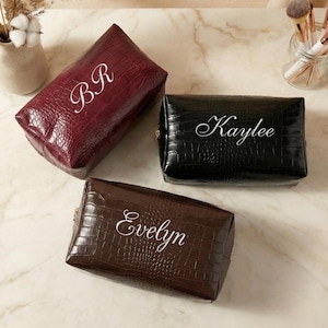 May include: Three personalised cosmetic bags in burgundy, black, and brown, each with a faux crocodile skin texture. The burgundy bag has the initials "BR", the black bag has the name "Kaylee", and the brown bag has the name "Evelyn" in white script.