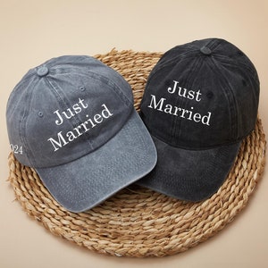 Może przedstawiać: Dwie szare i czarne czapki bejsbolowe z haftem "Just Married" w kolorze białym z przodu. Czapki leżą na plecionym słomkowym dywaniku.
