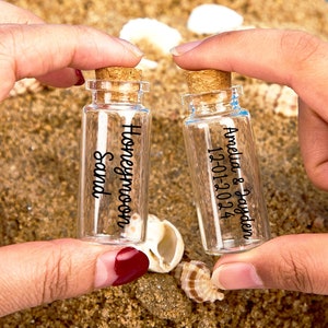 Pode incluir: Duas pequenas garrafas de vidro com rolhas de cortiça. Uma garrafa é etiquetada como "Honeymoon Sand" e a outra como "Amelia & Jayden 12-01-2024".