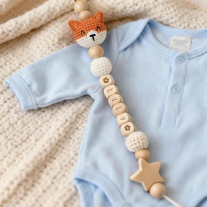 Op de afbeelding: Een baby fopspeenclip met een gehaakte vossenkop, houten kralen en de naam "LILY" op houten blokken. De clip heeft een houten ster en is bevestigd aan een lichtblauwe romper.